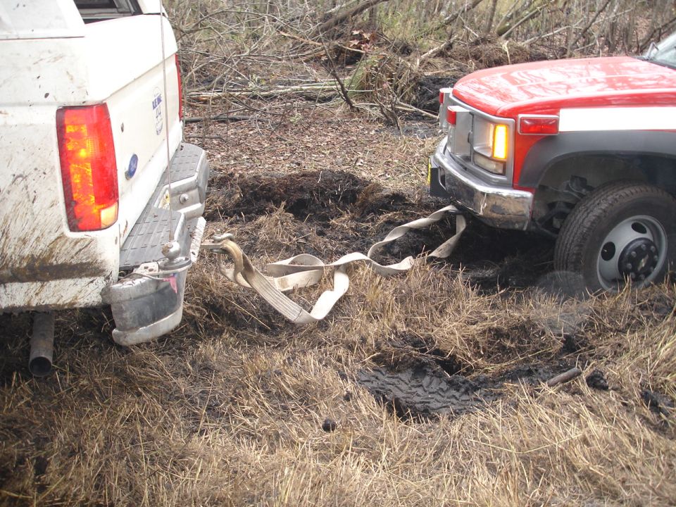 Rear view of a white 1989 Ford Bronco being towed by a red truck stuck in mud