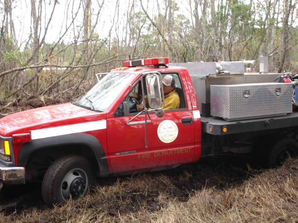 Red fire department truck with a white stripe on the side, driving through muddy terrain with trees in the background