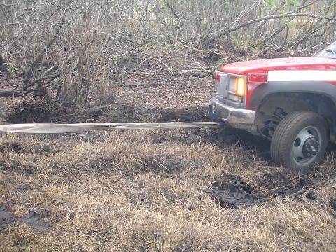 1989 Ford Bronco - A red truck is seen from the front, engaged in pulling a strap out of muddy, overgrown terrain.