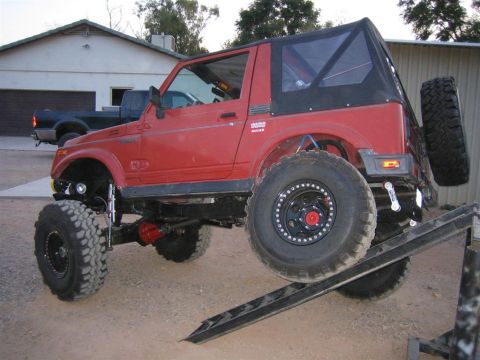 1987 Suzuki Samurai - A red 1987 Suzuki Samurai is shown with its front tire on a metal ramp, highlighting its off-road modifications.