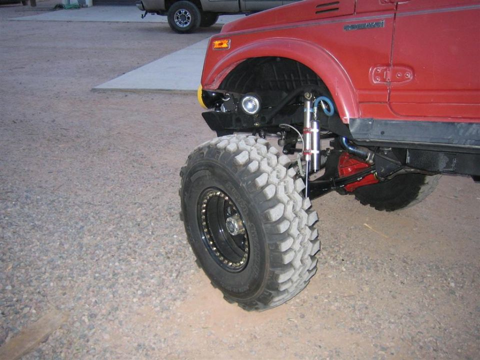 Front corner of a lifted red Suzuki Samurai showing remote-reservoir shock, red knuckle and mud tire
