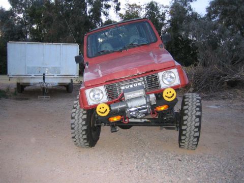 1987 Suzuki Samurai - A red 1987 Suzuki Samurai is parked on a dirt surface, equipped with a winch and distinctive smiley face auxiliary lights.