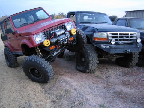 1987 Suzuki Samurai - Two off-road vehicles, a Suzuki Samurai and a Ford Bronco, are parked side-by-side on a dirt path.