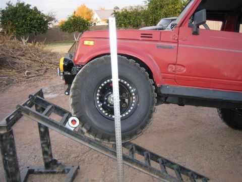 1987 Suzuki Samurai - A red off-road vehicle is positioned on a ramp with a ruler next to its large tire, likely for measurement purposes.