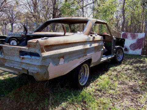 1964 Mercury Comet - A 1964 Mercury Comet body sits in a yard, showing significant rust and primer, with its engine and interior removed.