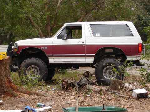 1989 Ford Bronco - This lifted 1989 Ford Bronco has a two-tone paint scheme and is parked in a wooded area with spare parts nearby.