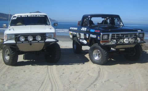 1990 Ford Bronco - white lifted truck and a black lifted Ford pickup with race number 300 parked side by side on a sandy beach.