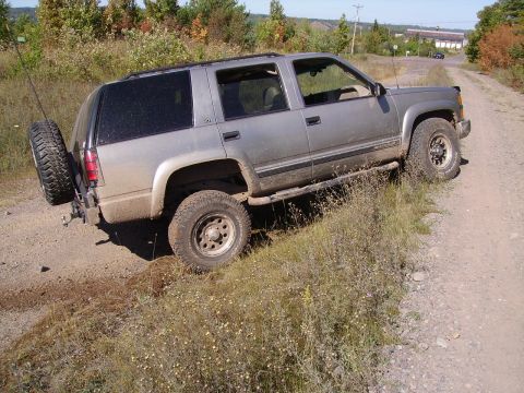 2000 Chevrolet Tahoe - SUV driving on a dirt road with overgrown grass on the side. Spare tire is mounted externally on the rear.