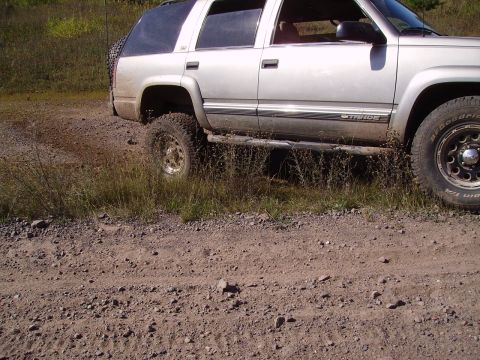 2000 Chevrolet Tahoe - Silver SUV with off-road tires parked on a dirt road, showing its side profile and a spare tire mounted on the rear.