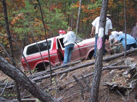 1995 Ford Bronco - A group of people are gathered around a red and white SUV that appears to be stuck on a wooded incline.