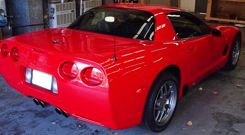 Red sports car, rear 3/4 view. Quad exhaust pipes, aftermarket wheels, visible antenna. Some scuffing on rear bumper