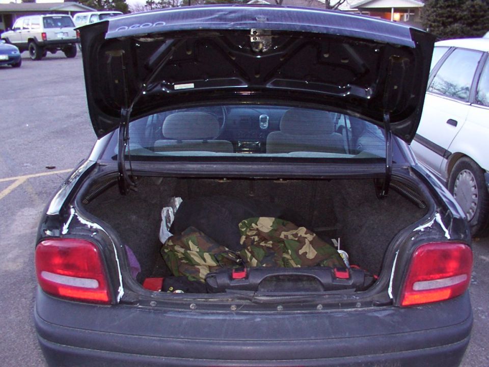 Black sedan trunk open, showing camouflage bags and a black case. White car parked to the right. Another SUV and car in background