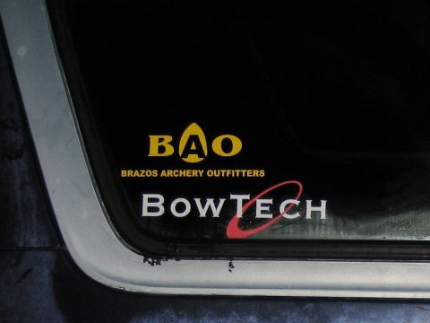 1986 Ford Bronco - Close-up of the lower corner of a truck's rear window carrying two vinyl decals: a yellow "BAO: Brazos Archery Outfitters" logo above a whi…