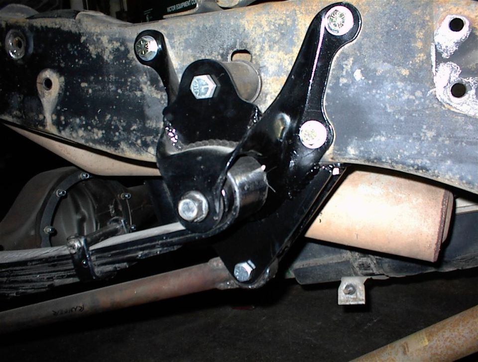 Black metal suspension bracket attached to a rusty frame with leaf springs and exhaust pipe visible
