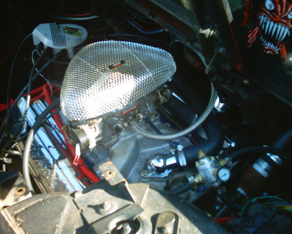 Engine bay with chrome teardrop air cleaner on carbureted V8, red plug wires, and fuel pressure gauge