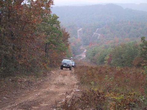 1995 Ford Bronco - Two SUVs navigate a winding dirt road through a wooded area with misty hills in the distance.