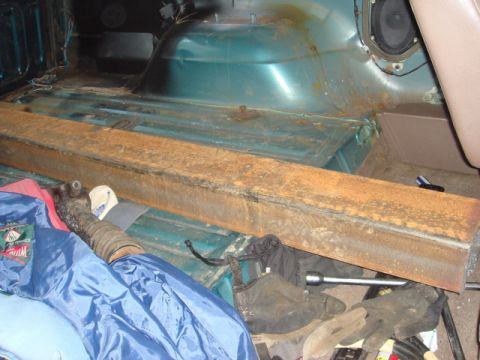 1996 Ford Bronco - rusty rectangular steel tube rests across a green vehicle interior floor near a wheel well speaker, with tools and a jacket on the ground.