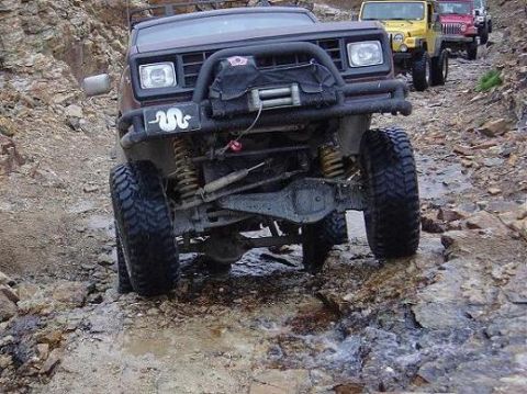 1986 Ford Bronco II - Modified 1986 Ford Bronco II navigates a rocky, wet trail with other vehicles following.