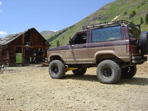 1986 Ford Bronco II - A lifted 1986 Ford Bronco II sits off-road near a dilapidated wooden building with mountains in the background.