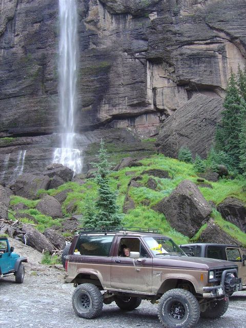 1986 Ford Bronco II - A lifted Ford Bronco II is parked in front of a tall waterfall in a rocky, green landscape.