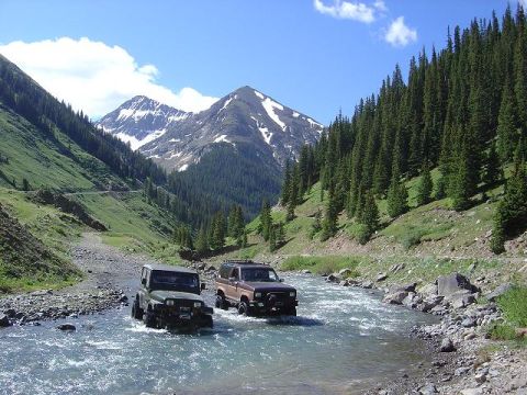 1986 Ford Bronco II - Two SUVs navigate a rocky riverbed surrounded by mountains and dense pine forests.