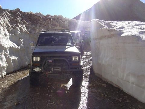 1986 Ford Bronco II - Several off-road vehicles navigate a path carved through high snowbanks under a bright sun.