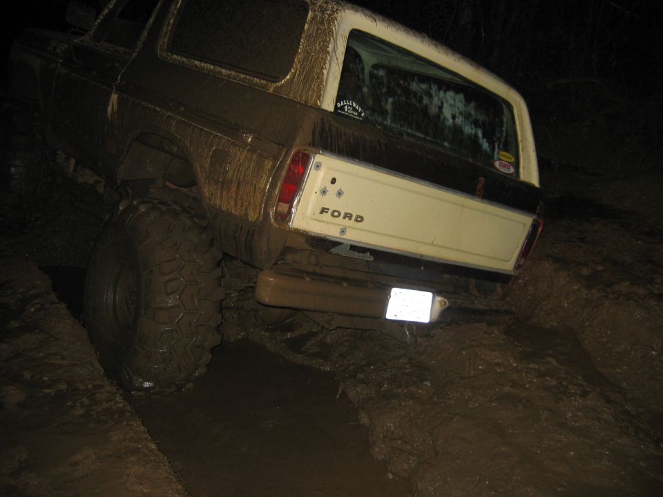 Mud-covered 1978 Ford Bronco with large tires driving through a muddy trail at night