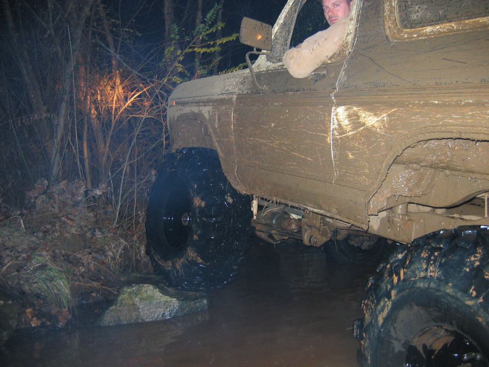 Mud-covered truck driving through water and rocks at night. Driver visible