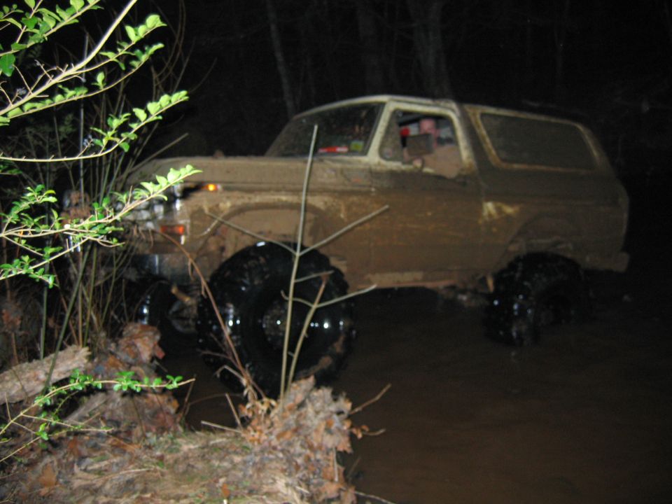 Mud-covered 1980 Ford Bronco driving through water, front 3/4 view