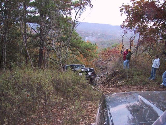 Two off-road vehicles are in a wooded area, one appears stuck and is being pulled by a rope. People are standing around