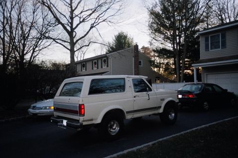 1989 Ford Bronco - A white Ford Bronco full-size SUV with a hardtop, tinted windows, red pinstripe and chrome wheels, parked in a driveway viewed from the rea…