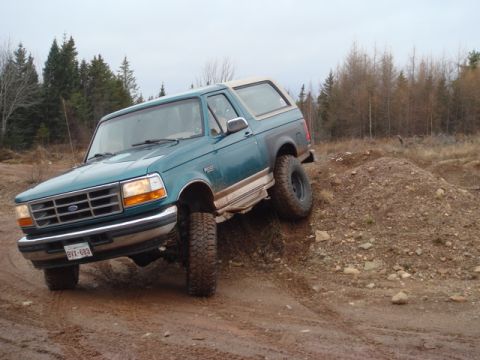 1996 Ford Bronco - A teal Ford Bronco navigates a muddy, off-road terrain with a pile of dirt to its right.