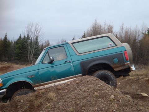 1996 Ford Bronco - This 1996 Ford Bronco navigates a challenging off-road terrain, its teal body and tan top clearly visible against the overcast sky.