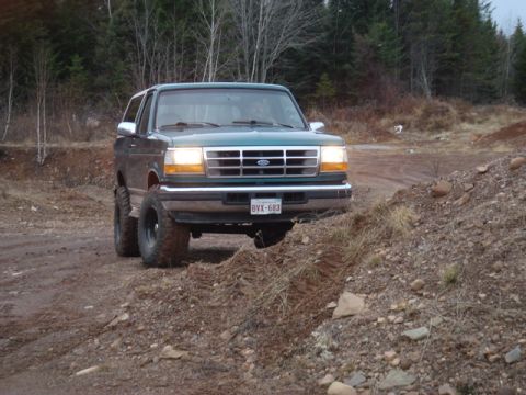 1996 Ford Bronco - A green Ford Bronco is shown driving on a dirt path with trees in the background.