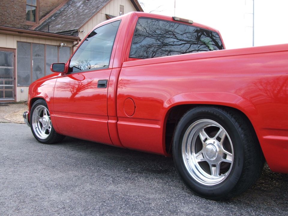 Red pickup truck parked on asphalt in front of a house. The truck has chrome wheels and a lowered stance