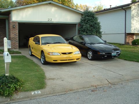 1998 Ford Mustang - Two cars, a yellow 1998 Ford Mustang and a black car, are parked in a residential driveway.
