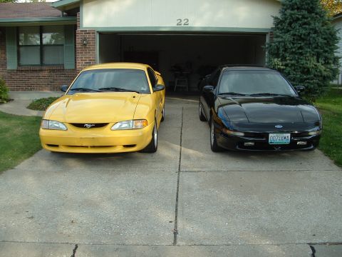 1998 Ford Mustang - A yellow Ford Mustang and a black Ford Probe sit side-by-side in a driveway in front of a garage.