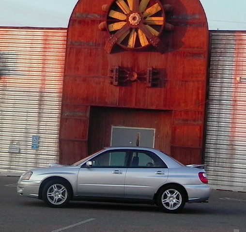 Silver sedan parked in front of a large rusty industrial structure with a fan