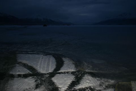 2003 Jeep TJ - Tire tracks are visible on an icy surface, leading towards a body of water with mountains in the background under a dark, twilight sky.