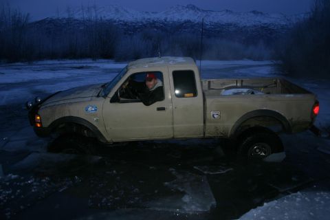 2003 Jeep TJ - A tan pickup truck is stuck in icy water with a snowy mountain range visible in the distance.