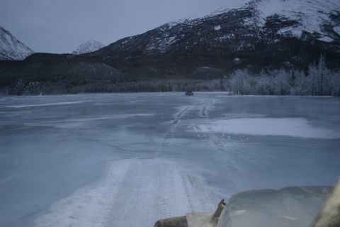 2003 Jeep TJ - An ATV traverses a frozen lake, leaving tracks in its wake under a cloudy sky with snow-capped mountains in the background.