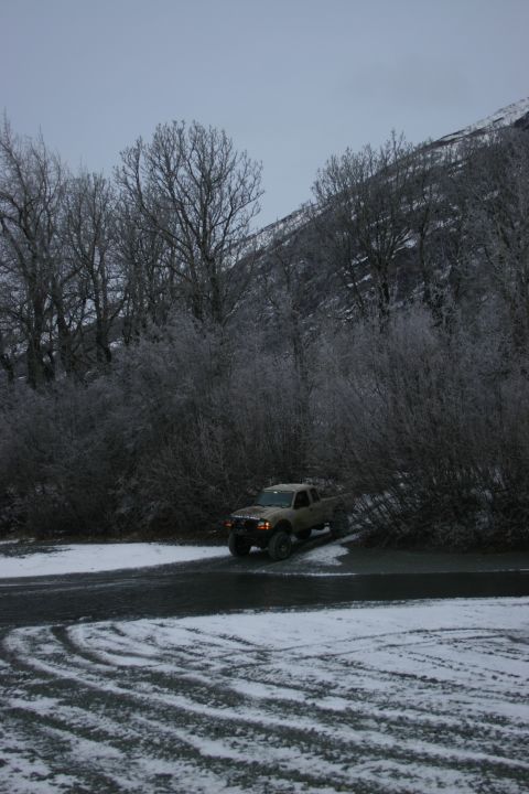 2003 Jeep TJ - A pickup truck drives on a gravel road covered in snow, with frosted trees and a mountain in the background.