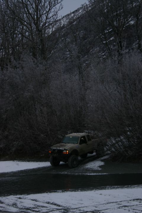 2003 Jeep TJ - A tan pickup truck drives on a snowy road surrounded by bare trees.