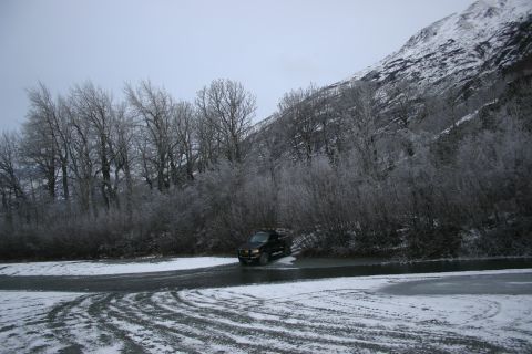 2003 Jeep TJ - A black pickup truck navigates a wet, snowy road with tire tracks visible, set against a backdrop of bare trees and a snow-dusted mountain.
