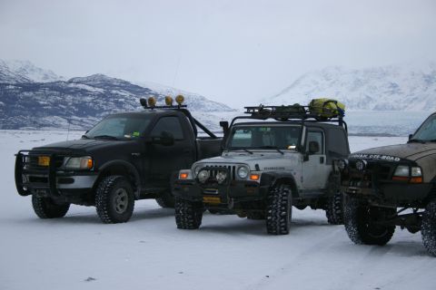 2003 Jeep TJ - Three modified off-road vehicles are parked in a snowy landscape with mountains in the background.