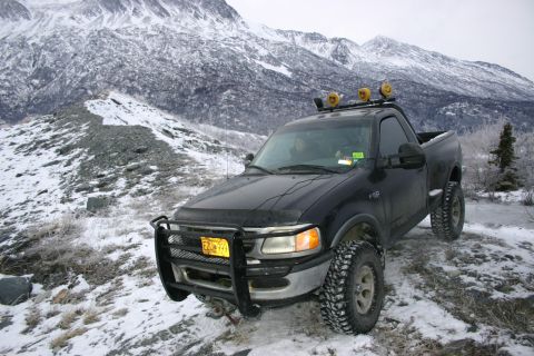 2003 Jeep TJ - A black Ford F-150 pickup truck is parked on a snowy mountain path with large mountains in the background.