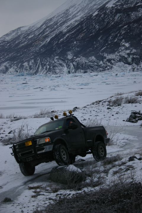 2003 Jeep TJ - A black pickup truck navigates a snowy and icy terrain with a large glacier visible in the background.