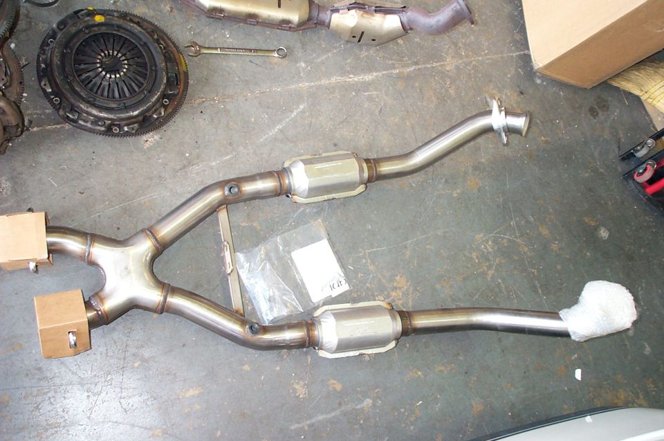 Stainless dual catalytic-converter X-pipe exhaust beside a clutch on shop floor