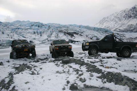 2003 Jeep TJ - Three modified off-road vehicles are positioned on a rocky, snow-covered area in front of a large glacier under an overcast sky.