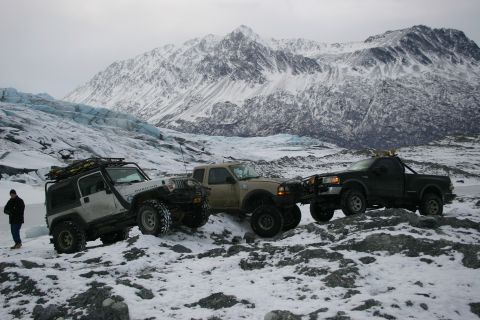 2003 Jeep TJ - Three modified trucks and SUVs are parked on a snowy landscape with a glacier and mountains behind them.